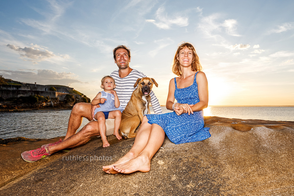 couple with baby and puppy sitting on rocks with sunset behind